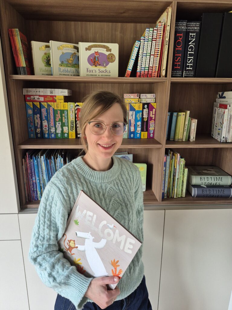 Nauczycielka języka angielskiego na tle regału z pomocami dydaktycznymi wypełnionego bogatą kolekcją materiałów do nauki języka angielskiego dla dzieci. Kolekcja zawiera popularne książeczki językowe, gry językowe i zestawy edukacyjne.